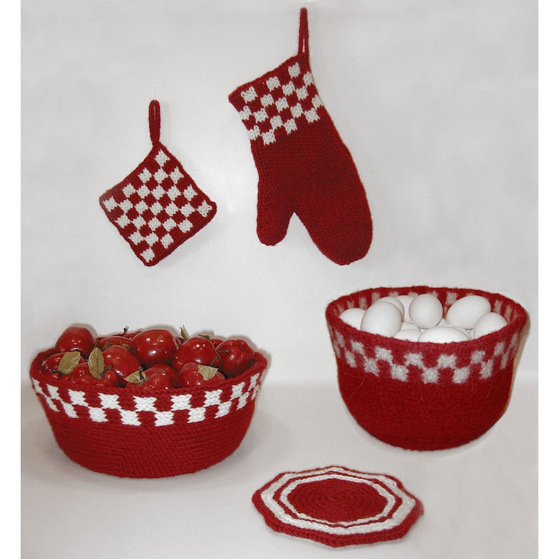 Op de afbeelding: Een verzameling rood-witte geblokte keukenaccessoires, waaronder een pannenlap, ovenwant, twee kommen en een onderzetter. E&eacute;n kom is gevuld met rode appels, de andere met witte eieren. De items zijn gebreid.