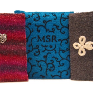 Op de afbeelding: Drie stoffen boekomslagen in rood, blauwgroen en bruin. De rode omslag heeft een zilveren hartvormige versiering. De blauwgroene omslag heeft zwarte krullen en de initialen "MSR". De bruine omslag heeft een cr&egrave;mekleurige bloemensluiting.