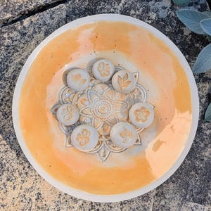 Könnte beinhalten: Ein runder Keramikteller mit glänzender orangefarbener und cremefarbener Glasur und einem zentralen geprägten Mandalamuster. Sieben kleine, kreisförmige, hellgraue Keramikplättchen mit goldfarbenen Pflanzen- und Insektenmotiven liegen darauf.