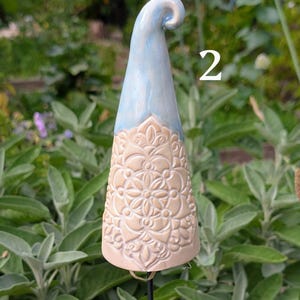 Könnte beinhalten: Ein konischer Gartenstecker aus Keramik mit einem hellblau glasierten oberen Teil, der sich zu einer Spitze krümmt, und einer beigen, unglasierten Basis, die mit einem komplizierten, geprägten Blumen-Mandala-Muster verziert ist. Die Zahl 2 ist in weißem Text sichtbar.