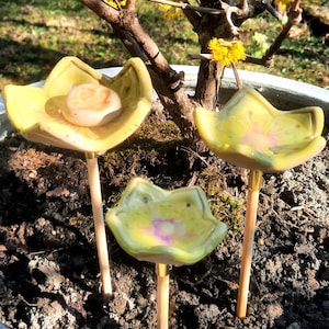Könnte beinhalten: Drei Gartenstecker aus Keramik in Blumenform in einem Topf. Die Stecker sind gelb mit einem hellrosa Zentrum und an Holzstäben befestigt. Der Topf ist mit dunkler Erde gefüllt und steht neben einem blühenden Baum mit gelben Blüten.
