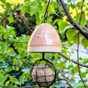 Könnte beinhalten: Ein hängendes Vogelfutterhaus mit einer glatten, gesprenkelten hellrosa Keramikkuppel und einem darunter liegenden schwarzen Drahtkäfig. Der Käfig enthält eine runde, texturierte, beigefarbene Fettkugel. Alles hängt an einem schwarzen Metallhaken.