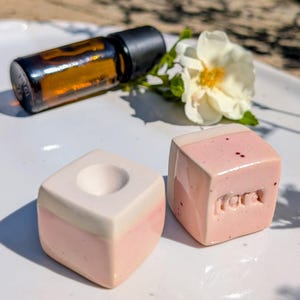 Könnte beinhalten: Zwei Keramikwürfel mit einer rosa und weiß glasierten Oberfläche. Ein Würfel hat eine kreisförmige Vertiefung auf der Oberseite, während der andere den geprägten Schriftzug "flora" an der Seite aufweist. Eine braune Glastropfflasche und eine weiße Blume liegen daneben.