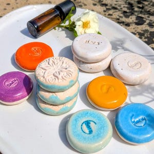 Könnte beinhalten: Eine Sammlung runder Keramikscheiben in verschiedenen Farben wie Orange, Lila, Gelb, Blau und Weiß, teilweise mit geprägten Blumenmustern oder Text. Eine kleine bernsteinfarbene Glasflasche und eine weiße Blume liegen zwischen den Keramikstücken.