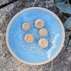 Könnte beinhalten: Eine runde, hellblaue Keramikschale mit einem geprägten Mandalamuster auf der Oberfläche. Fünf kleine, kreisförmige, cremeweiße Keramikplättchen mit goldfarbenen Pflanzen- und Insektenmotiven sind in der Mitte der Schale angeordnet.