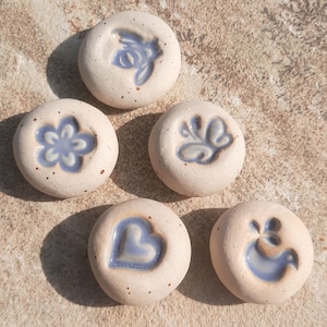 Könnte beinhalten: Sechs kleine, runde Keramiksteine. Jeder Stein hat eine hellbeige Grundfarbe mit einem blauen Design. Die Designs umfassen eine Blume, ein Herz, einen Schmetterling und einen Vogel. Die Steine sind auf einer strukturierten Oberfläche angeordnet.