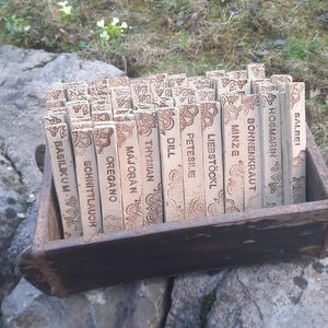 Könnte beinhalten: Eine Holzkiste gefüllt mit rechteckigen Kräutermarkern. Jeder Marker ist hellbraun mit einem dekorativen Muster und dem Namen eines Krauts in dunkelbraunem Text. Die Kiste steht auf einem großen Stein, mit Gras und kleinen Blumen im Hintergrund.