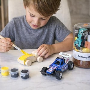 May include: A child paints a wooden toy car yellow with a paintbrush. Beside the car are small paint pots in red, blue, yellow, and black. A blue and white toy race car and a sensory play jar are also on the table.