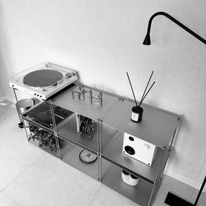 May include: A modern, minimalist metal shelving unit with a turntable, speaker, and decorative items. The unit has multiple shelves and a sleek, industrial design. A black floor lamp is in the background.