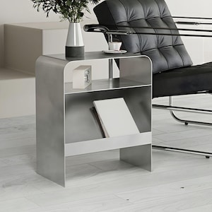 Può includere: Un tavolino moderno in metallo spazzolato con due ripiani. Un vaso bianco e grigio con vegetazione, un piccolo blocco bianco e una tazza di caffè sono sopra. Una sedia in pelle nera è sullo sfondo. Il tavolo ha un design elegante e minimalista.