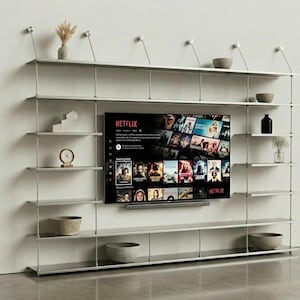 Op de afbeelding: Een grote, aan de muur gemonteerde metalen stellingkast met meerdere horizontale niveaus en dunne verticale steunen. Een flatscreen-tv is in het midden gemonteerd en toont de Netflix-interface. Diverse decoratieve kommen en objecten staan op de planken.