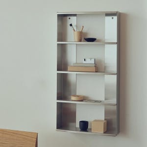 Può includere: Una mensola a parete in acciaio inossidabile con quattro scomparti. I ripiani contengono vari oggetti, tra cui una tazza di legno con pennelli, una piccola ciotola nera, libri, un disco di legno e una tazza nera. È presente anche una piccola scatola marrone chiaro.