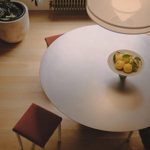 May include: A large round table with a smooth surface, accompanied by two small stools with dark red corduroy seats and light-coloured legs. A shallow bowl filled with yellow lemons and green leaves sits in the centre of the table.