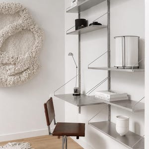 May include: A wall-mounted metal shelving system with multiple tiers and an integrated desk surface. A brown leather chair with a metal frame sits in front of the desk. The shelves hold various white and black decorative objects and books.