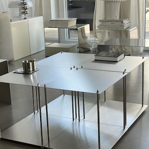 May include: A contemporary, silver metal coffee table with a square design. The table has a flat top and base, supported by thin, cylindrical legs. A cup and saucer sit on the table's surface. The room also features white furniture and a lamp.