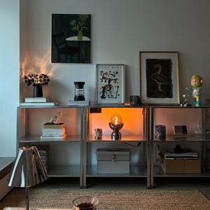 May include: A modular metal shelving unit featuring three sections filled with various decorative objects, including a black lamp, framed art prints by Joan Miro, a small figurine, a glowing orange orb lamp, and a silver table lamp in the foreground.