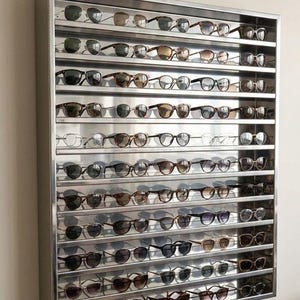 Op de afbeelding: Een rechthoekige wandvitrine met een zilverkleurig metalen frame en meerdere horizontale planken. De vitrine is gevuld met talloze zonnebrillen en brillen in verschillende stijlen, kleuren en montuurvormen, netjes gerangschikt in rijen.