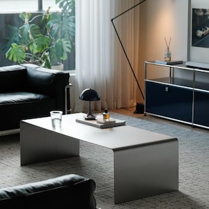 Op de afbeelding: Een matgrijze metalen salontafel met een doorlopend watervalrandontwerp. Het rechthoekige oppervlak bevat een kleine zwarte koepelvormige lamp, een helder glas water en een klein glazen flesje met een kurk op een plat dienblad.