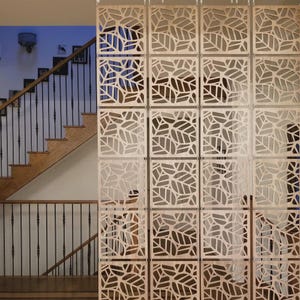 Può includere: Un divisorio composto da diversi pannelli quadrati in legno chiaro collegati da piccoli ganci metallici. Ogni pannello presenta un motivo tagliato al laser di foglie stilizzate con venature complesse, creando un design a griglia geometrica.