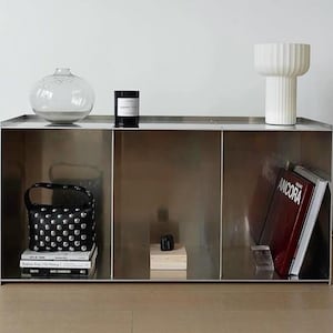Puede incluir: Un moderno mueble metálico con tres compartimentos. Presenta un jarrón de cristal transparente, una vela negra, un bolso negro con tachuelas, libros, una lámpara blanca y un libro rojo titulado "ANCORA". El mueble está en una habitación de tonos neutros.