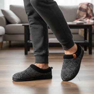 Op de afbeelding: Grijze en zwarte gebreide pantoffels met een zwarte zool en bies. De pantoffels hebben een zachte, donzige voering en een klein bruin label aan de zijkant. De afbeelding toont de pantoffels op een houten vloer.