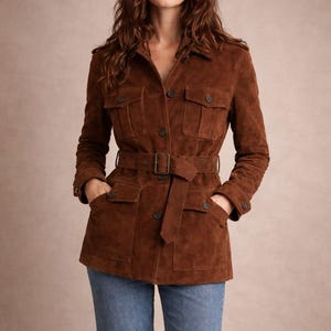 May include: A brown suede jacket with a belted waist and multiple pockets. The jacket has a button-up front and a classic, tailored design. The model is wearing blue jeans. The jacket is a versatile outerwear piece.