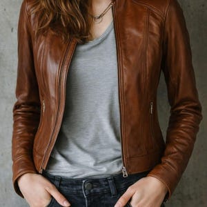 May include: A brown leather jacket with a stand-up collar and a front zipper. The jacket has two zippered pockets and is worn over a gray t-shirt and dark wash jeans. The model has their hands in their pockets.