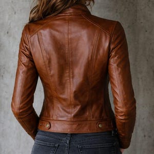 May include: A brown leather jacket with a classic design. The jacket features a stand-up collar, long sleeves, and button details at the waist. The model is wearing dark wash jeans.