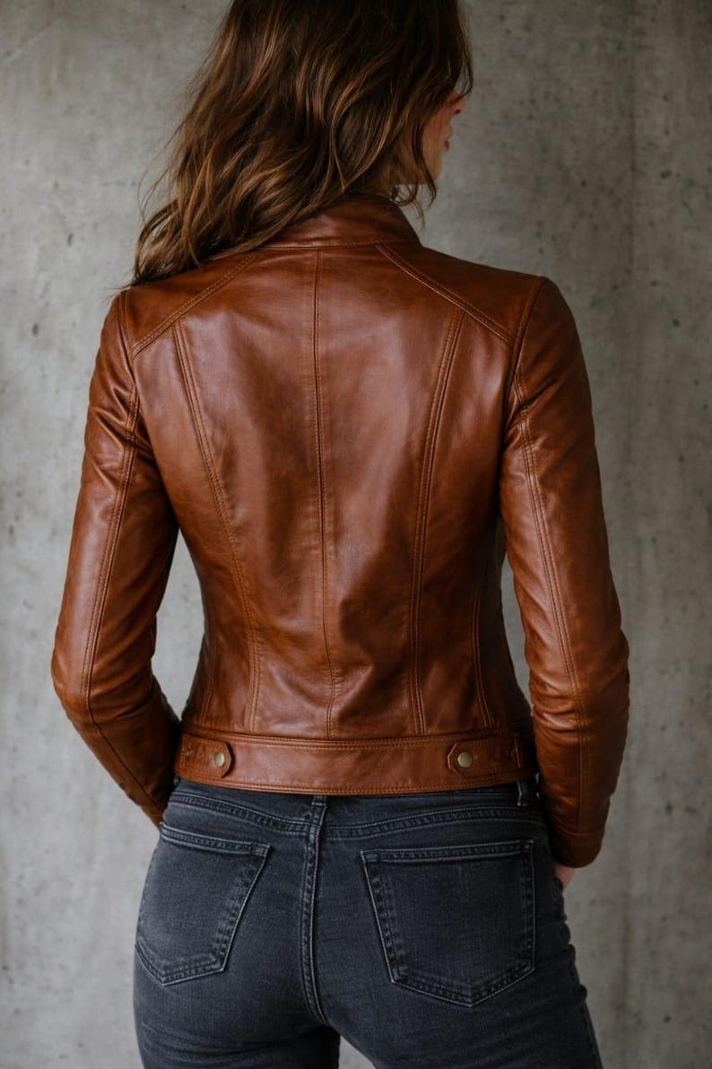 May include: A brown leather jacket with a classic design. The jacket features a stand-up collar, long sleeves, and button details at the waist. The model is wearing dark wash jeans.