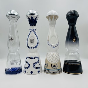 Op de afbeelding: Vier verschillende tequilaflessen met unieke ontwerpen. De eerste is van helder glas met een zilveren dop. De tweede is van wit keramiek met blauwe bloemmotieven. De derde is beige en wit met blauwe accenten. De vierde is een zwart-heldere fles.