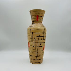 Op de afbeelding: Een hoge, beige keramische vaas met een modern design uit het midden van de eeuw. De vaas heeft zwarte, golvende lijnen en rode rechthoekige accenten. De vaas heeft een brede basis die taps toeloopt naar een smallere hals en bovenaan uitloopt.