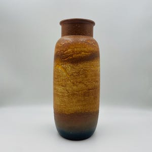 Op de afbeelding: Een hoge, cilindrische keramische vaas met een getextureerd oppervlak. De vaas heeft een kleurengradiënt, van een donkerbruine basis naar tinten beige en bruin, met een donkerdere bruine rand. Klassiek, tijdloos ontwerp.