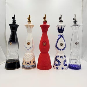May include: Five tall, slender glass bottles with pourer spouts. From left to right: a black gradient bottle, a cream bottle with blue and gold floral patterns, a solid matte red bottle, a white bottle with blue floral designs, and a clear glass bottle.