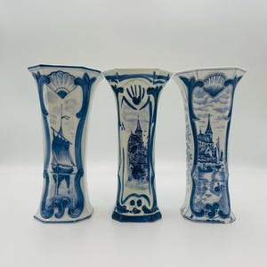 Może przedstawiać: Trzy biało-niebieskie ceramiczne wazony ze szczegółowymi wzorami marynistycznymi i architektonicznymi. Każdy wazon przedstawia unikalną ilustrację żaglówki, zamku i nadmorskiego miasteczka. Wazony mają rozkloszowaną górę i szczegółową podstawę.