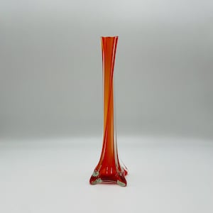 Peut inclure: Un vase en verre haut et élancé présentant une base transparente qui se transforme en un motif en spirale orange vif. Le vase possède un pied évasé à quatre pointes et un col cylindrique étroit avec une finition lisse et polie.