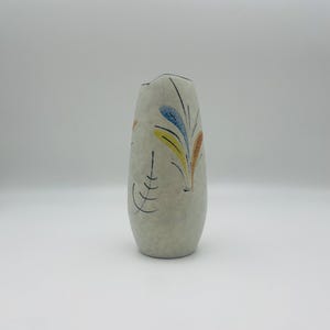 Op de afbeelding: Een taps toelopende keramische vaas met een matte gebroken witte afwerking, voorzien van handgeschilderde abstracte bladmotieven in blauw, geel en oranje, geaccentueerd door dunne zwarte lijnen rond het gebogen lichaam.