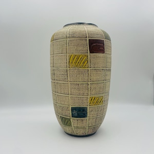 Op de afbeelding: Een hoge, beige keramische vaas met een rasterpatroon en kleurrijke vierkante accenten. De vierkanten hebben ontwerpen in rood, geel, groen en donkerblauw. De vaas heeft een donkerblauwe rand en een getextureerd oppervlak.