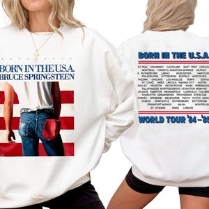 May include: White sweatshirt featuring the text "BORN IN THE U.S.A. BRUCE SPRINGSTEEN" above an image of a person in jeans and a red pocket. The back of the sweatshirt lists tour cities and "WORLD TOUR '84-'85".