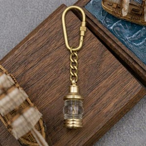 May include: A brass-toned metal keychain featuring an elongated oval carabiner clip with a screw-lock mechanism, a short chain link, and a miniature lantern-shaped pendant with a clear glass center and metallic ribbed top and bottom sections.