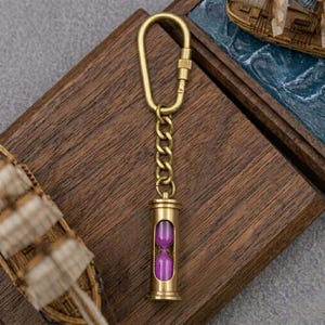 May include: A brass keychain featuring a carabiner clip attached to a short chain and a small cylindrical hourglass pendant. The hourglass has a metallic brass frame and contains vibrant purple sand, visible through the clear glass center.