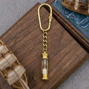 May include: A brass-toned metal keychain featuring a carabiner clip attached to a short chain and a miniature hourglass pendant. The hourglass has a clear glass center filled with white sand, encased in a cylindrical brass-colored metal frame.
