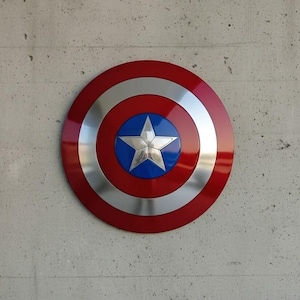 May include: A round shield with a red outer ring, a silver inner ring, and a blue center. A silver five-pointed star is in the center. The shield is mounted on a gray concrete wall.