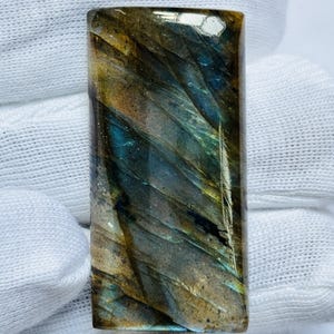Puede incluir: Una piedra preciosa rectangular de labradorita con una superficie pulida. La piedra muestra una mezcla de colores, incluyendo azules, verdes y dorados, con un efecto brillante e iridiscente. El patrón de la piedra tiene una apariencia natural y en capas.