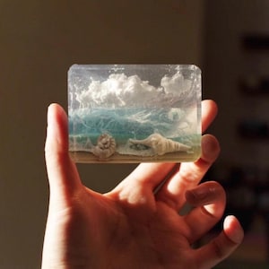 May include: A clear, rectangular resin block featuring a miniature beach scene. The scene includes a sandy shore, seashells, blue ocean waves, and fluffy white clouds. The block is held in a hand.
