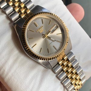 May include: A Seiko 5 automatic wristwatch featuring a silver sunray dial, gold-tone fluted bezel, and a two-tone stainless steel jubilee-style bracelet with gold center links and silver outer links. The dial displays the Seiko logo and day-date.