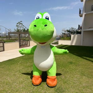 May include: A full-body costume of a green dinosaur-like character with a large head, white belly, and orange boots. The character has large, expressive eyes and a friendly smile. The costume is standing on green grass under a blue sky.