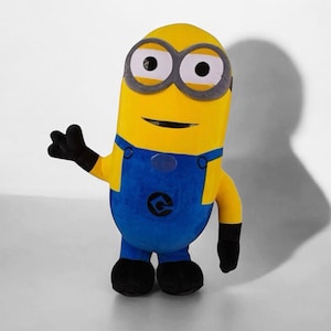 May include: A plush toy shaped like a yellow cylindrical character with two large white eyes framed by grey circular goggles. The figure wears blue overalls with black buttons and a black logo on the front, along with black gloves and black shoes.