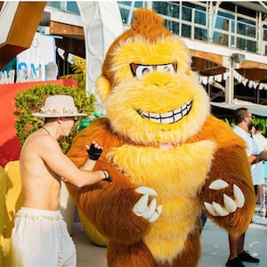Puede incluir: Una persona interactúa con un gran disfraz de gorila peludo. El disfraz es principalmente amarillo y marrón, con una cara sonriente y grandes manos blancas. La persona lleva un sombrero y pantalones blancos. La escena tiene lugar al aire libre.