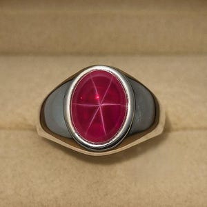 May include: A silver ring with an oval, star ruby gemstone. The ruby is a deep red color with a six-ray star pattern. The ring is set in a polished silver band and is displayed in a brown jewelry box.
