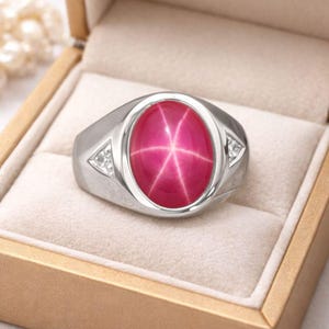 May include: A silver ring with a large, oval, ruby-colored gemstone. The gemstone has a star-like pattern. Small, clear stones are set on either side of the main stone. The ring is presented in a beige jewelry box.